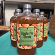 Load image into Gallery viewer, A clear glass bottle of Apple Pie Shrub with a green label and black cap, standing on a teal tablecloth with several matching bottles behind it under a white canopy tent.
