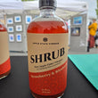 Load image into Gallery viewer, A bottle of Apple State Vinegar Strawberry &amp; Rhubarb Shrub, featuring a beige and red label, set on a black table with an outdoor market background.
